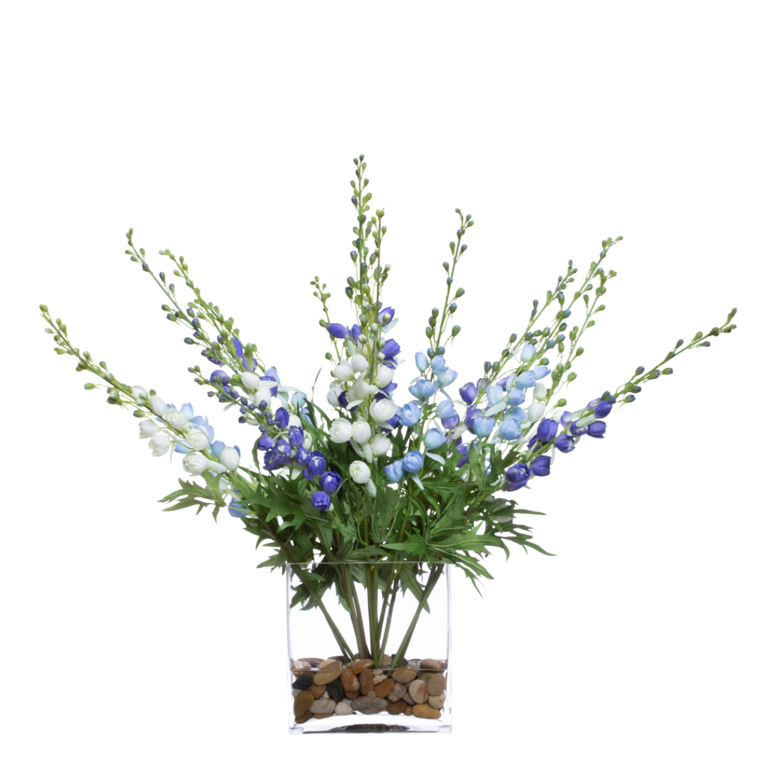 Artificial delphinium flower arrangement with blue and white blooms in a glass vase with stones