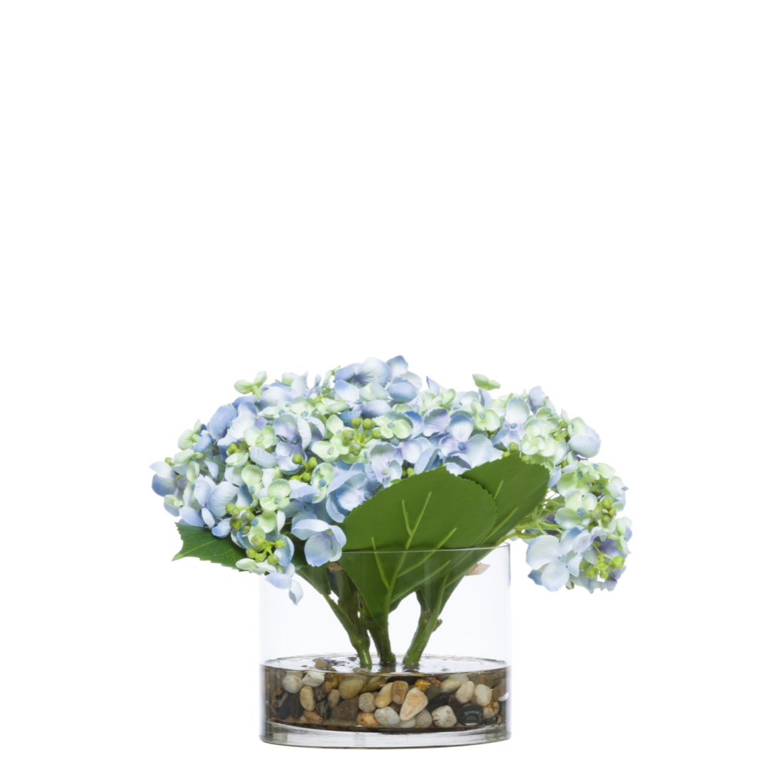 Artificial blue hydrangea floral arrangement in glass vase with pebbles, white background