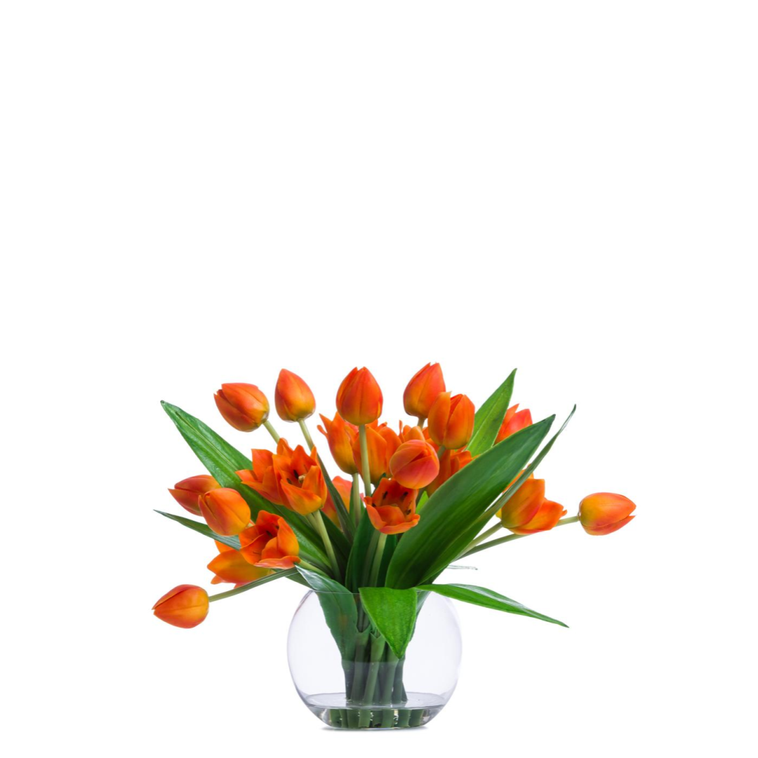 Vibrant orange artificial tulip arrangement in clear round glass vase with green leaves