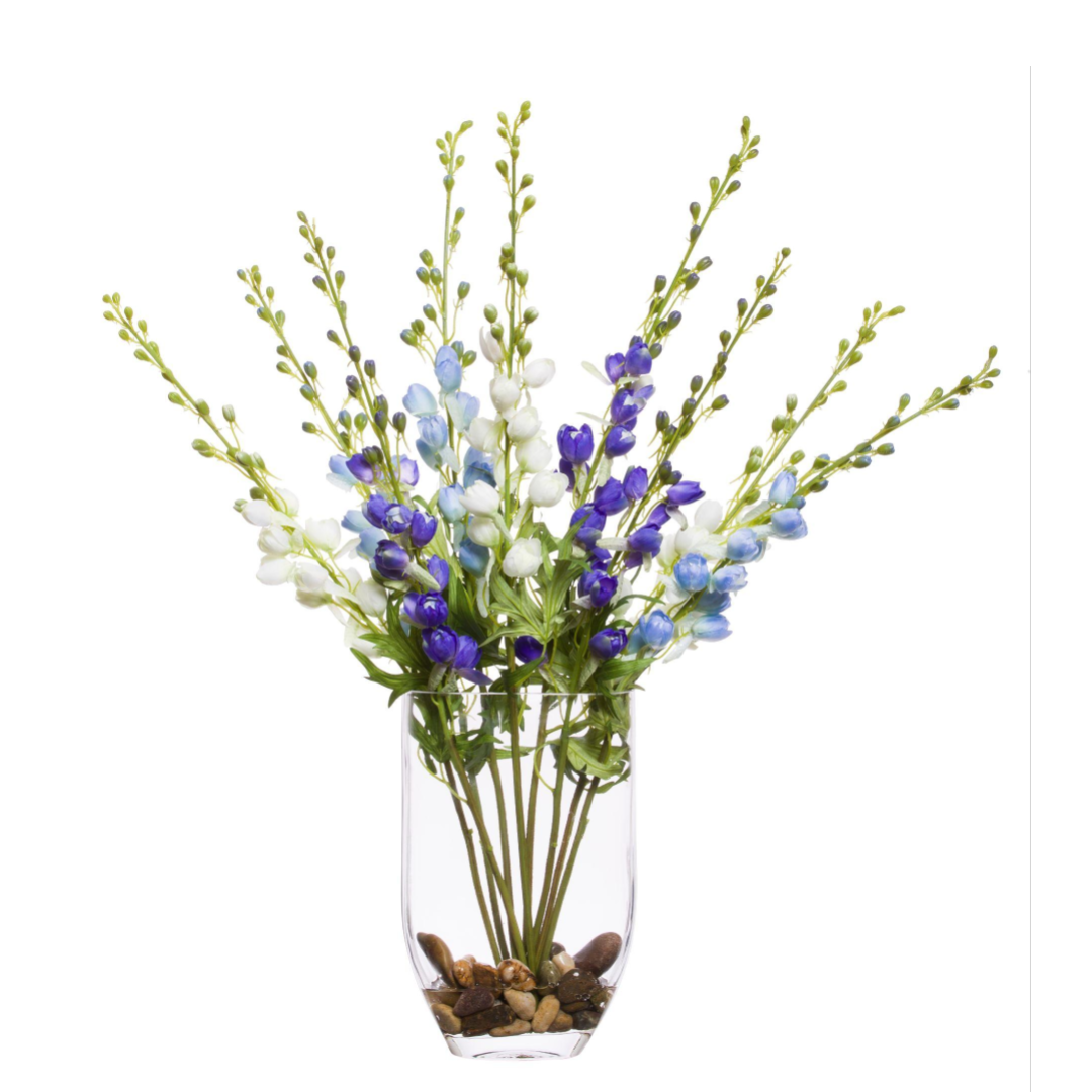 Artificial blue and white delphinium flowers in a clear glass vase with river stones