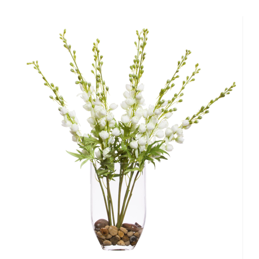 White artificial delphinium flowers in a clear vase with pebbles, isolated on white background.