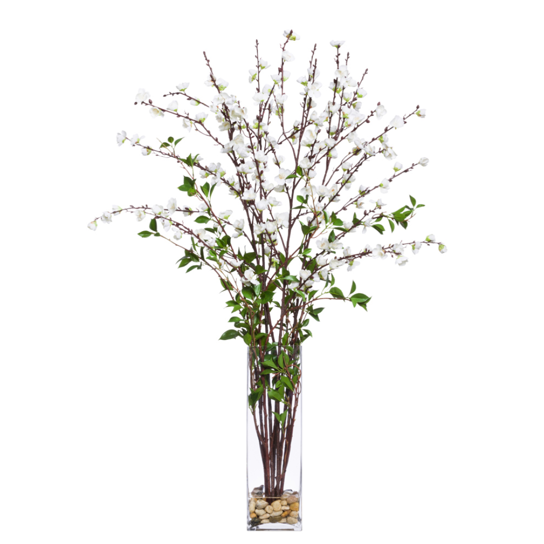 Artificial plum blossom branches with white flowers in a tall glass vase with stones.