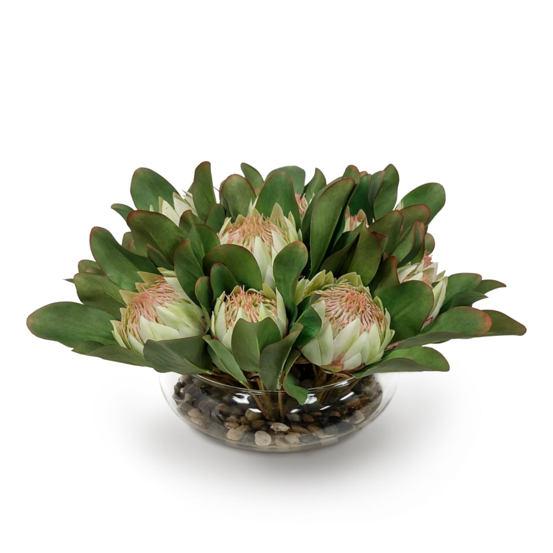 Artificial protea flower arrangement in clear glass bowl with pebbles on white background