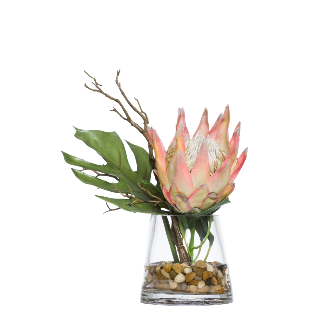 Artificial protea flower arrangement with split leaves in a clear vase with stones